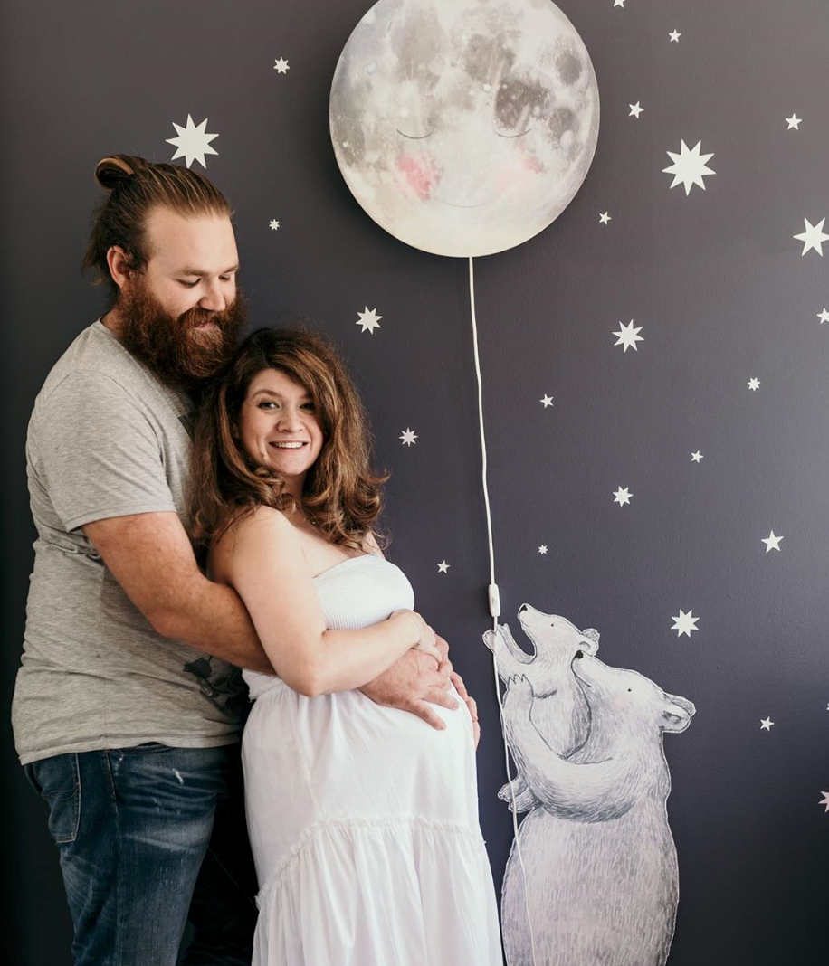Werdende Eltern neben der Schlafender-Mond-Wandlampe mit weißen Sternenstickern fürs Babyzimmer